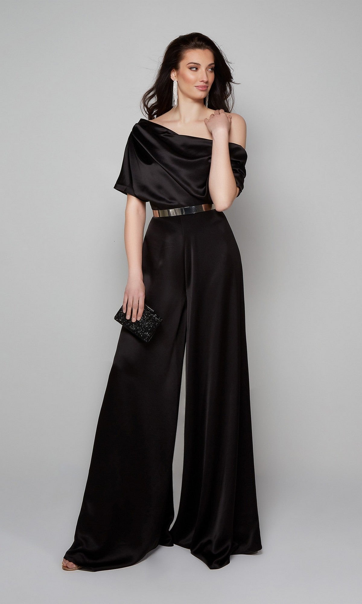 A woman models the Alyce 70019 long formal jumpsuit in black, featuring an off-shoulder neckline and wide-leg pants, paired with a metallic belt, long earrings, and a black clutch against a plain background.